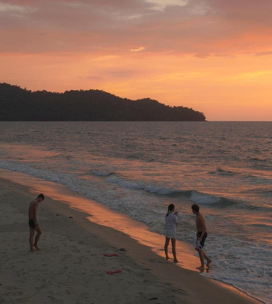 A serene beach scene at sunset, featuring gentle waves lapping against a sandy shore. Silhouettes of three individuals are seen enjoying the calm waters. The horizon showcases a blend of warm colors reflecting off the water, with a backdrop of distant hills.