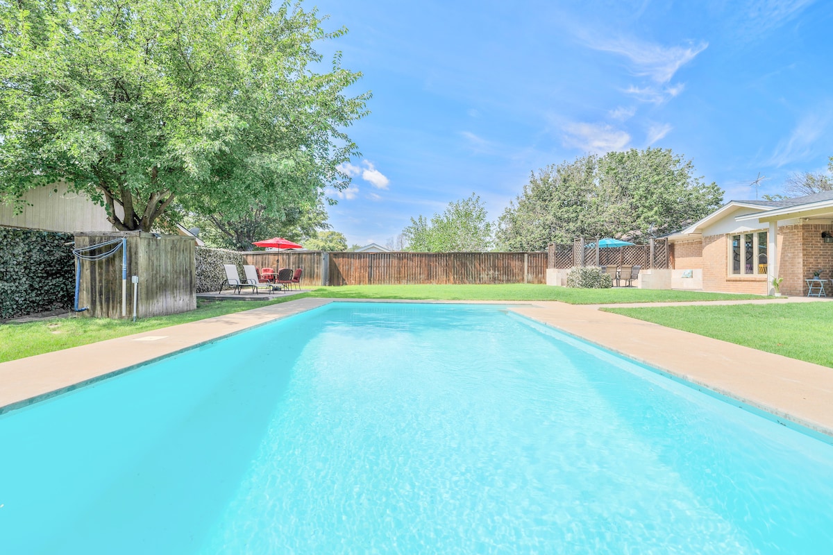 The image presents a clear blue swimming pool surrounded by a spacious grassy area. Lush trees provide shade, with lounge chairs positioned nearby. Outdoor seating and an umbrella are visible in the background, enhancing the inviting outdoor space.