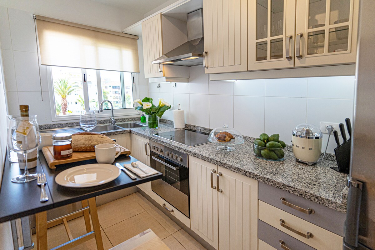 A modern kitchen is presented with light-coloured cabinetry and granite countertops. A variety of kitchen appliances, including a blender and a stove, are visible. Natural light enters through a window, illuminating the countertop where a loaf of bread and a bowl of limes are arranged.