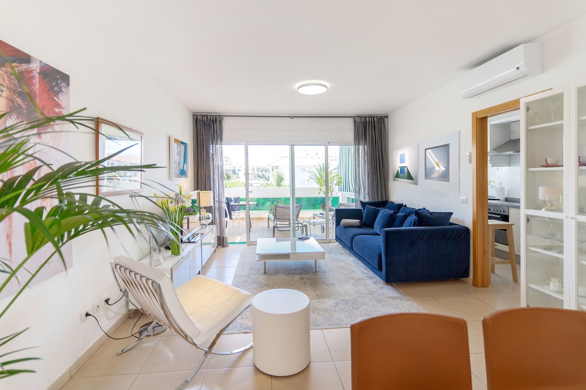 A spacious living area features a comfortable blue sofa, a white coffee table, and a modern armchair, creating an inviting atmosphere. Large sliding glass doors allow natural light to fill the room, with access to a terrace visible in the background.