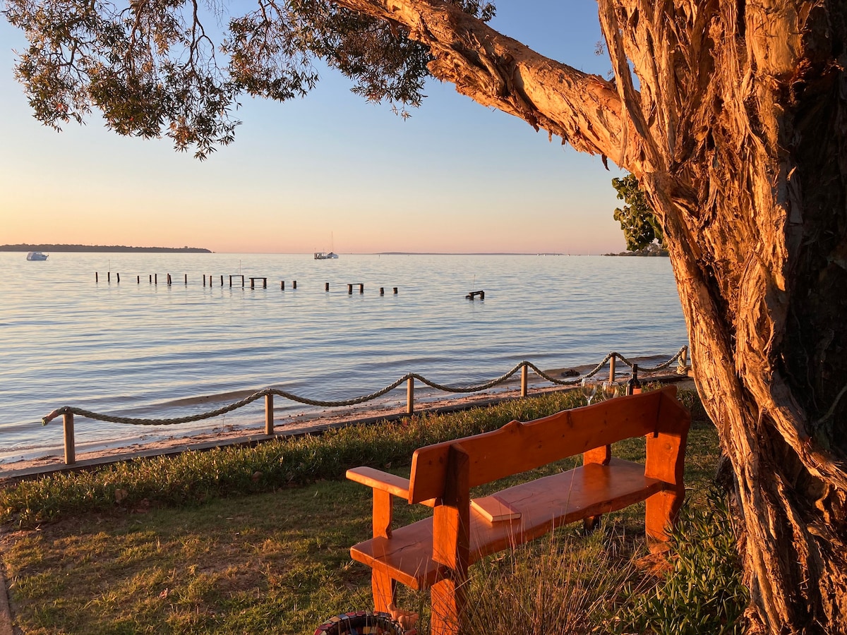 A wooden bench is positioned under a tree, offering a view of calm waters at sunset. Silhouetted against the horizon, a few boats can be seen in the distance, while old wooden posts rise from the water, adding to the tranquil scene.