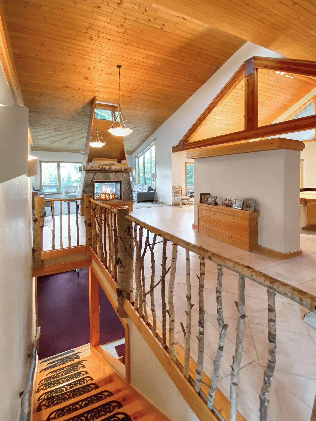 A spacious interior is highlighted by high wooden ceilings and large windows allowing natural light to flow in. An inviting staircase with a railing made of birch logs leads to a sunlit living area, while a warm glow emanates from a fireplace visible in the distance.