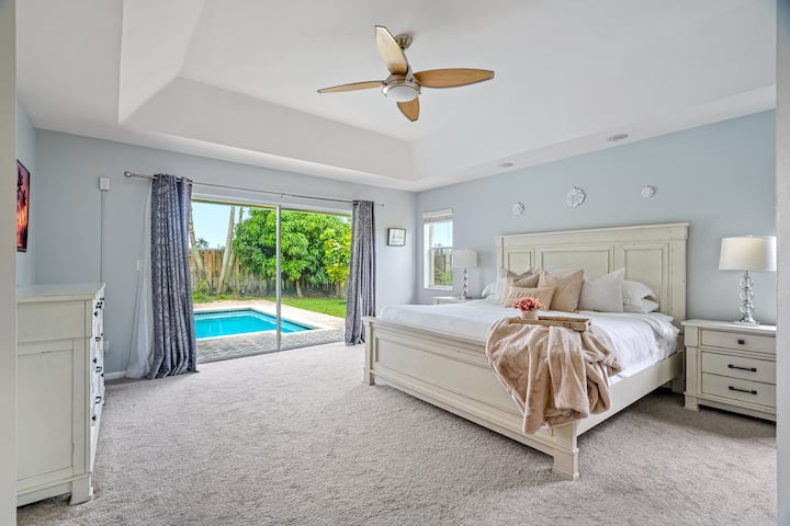 Primary Bedroom with king bed, ceiling fan, ensuite bathroom with separate tub and shower and views of the heated pool and tropical landscaping. Fort Lauderdale Area Pool Home.