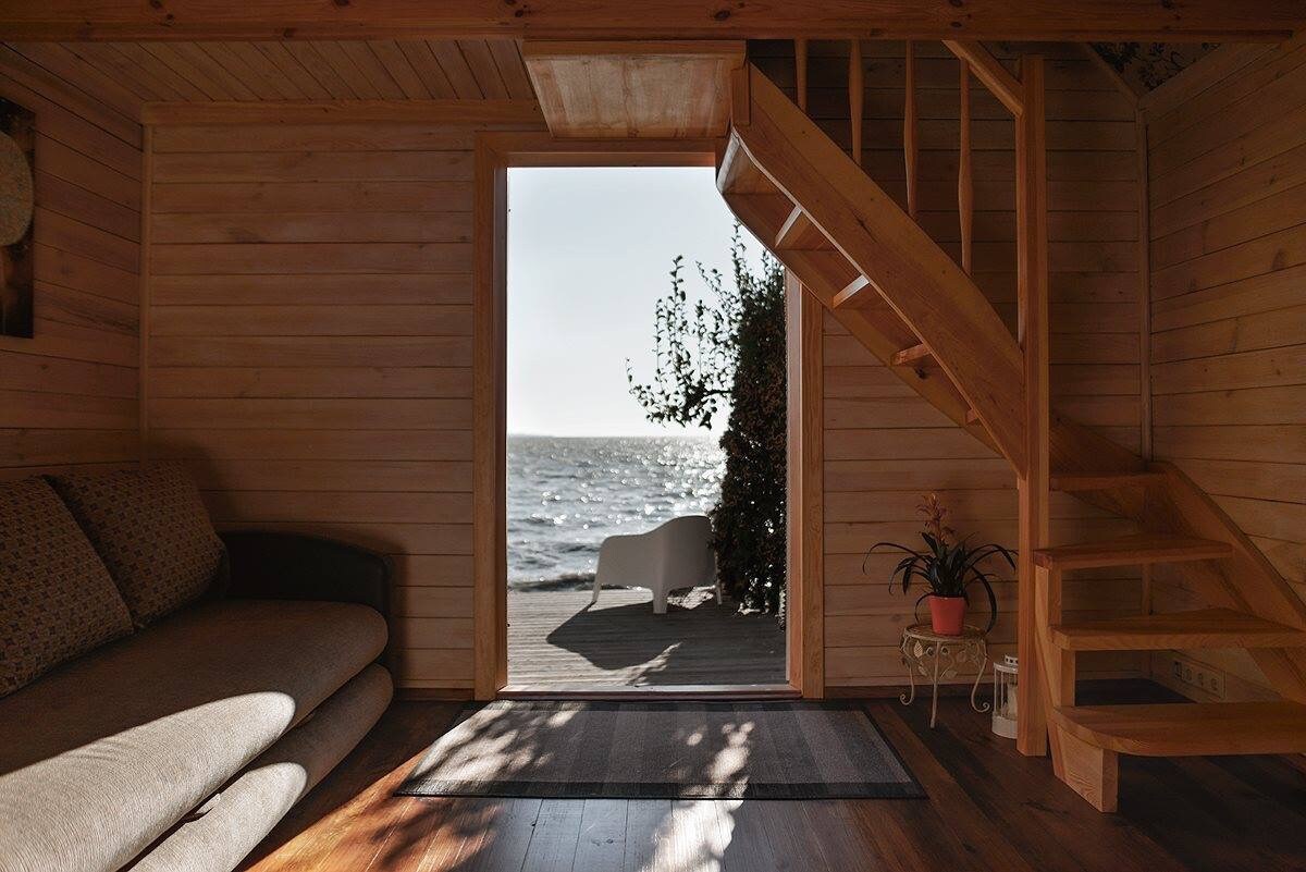 The interior features warm wooden walls and a staircase leading to an upper level. A large opening reveals a view of the shimmering water outside, with a white lounge chair positioned on the deck. Natural light fills the space, enhancing the inviting atmosphere.