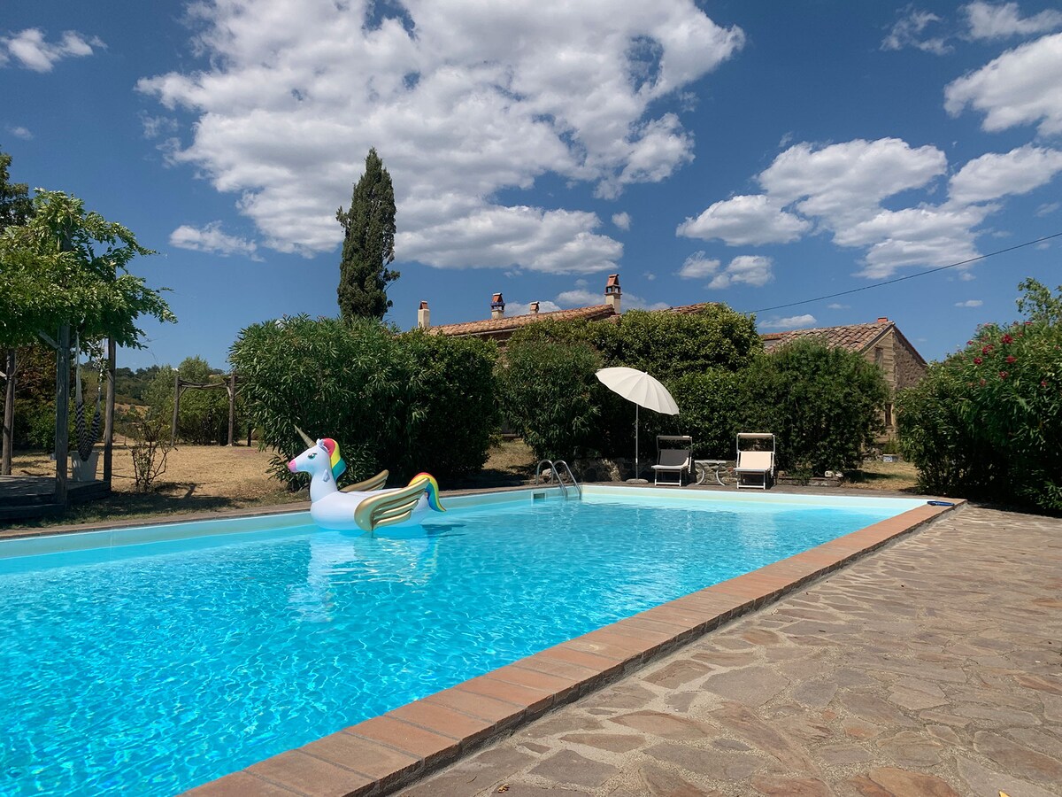 A serene swimming pool is framed by lush greenery and a sun terrace. A unicorn-shaped inflatable floats on the clear water under a bright sky dotted with clouds, while loungers and a shaded umbrella provide relaxation spaces nearby.
