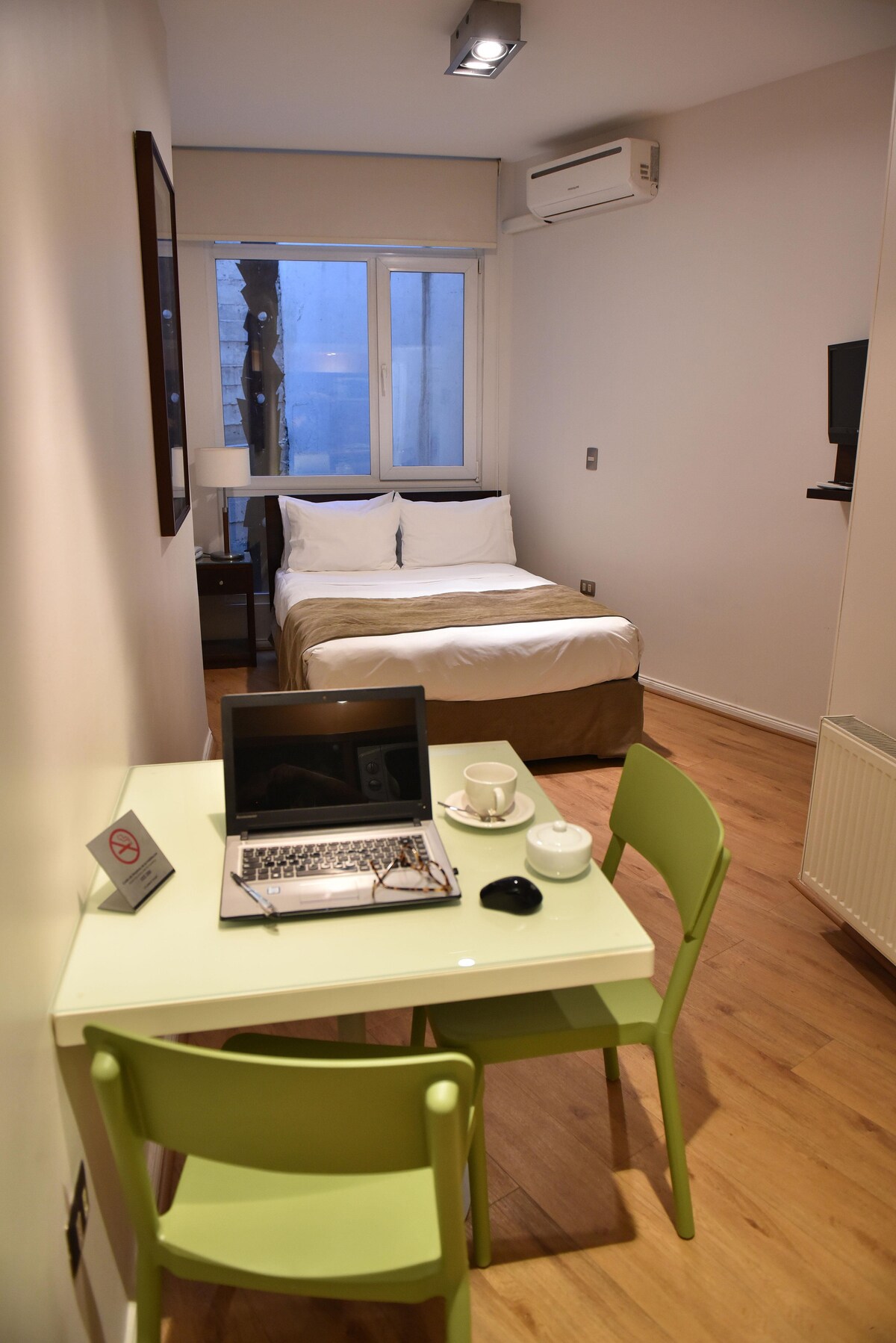 A spacious room features a comfortable bed with light-colored bedding, complemented by a window allowing natural light. A small green table with two matching chairs is arranged beside a laptop and cup set, providing a functional work area. Modern furnishings enhance the overall decor.