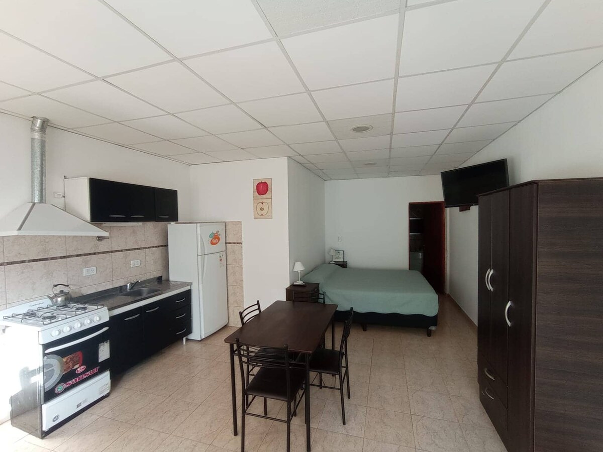 A spacious room contains a king-size bed and a single bed. A small dining table with four chairs is positioned nearby. The kitchen area features a gas stove, refrigerator, and cabinetry. Light-colored tiles cover the floor, contributing to a bright ambiance.