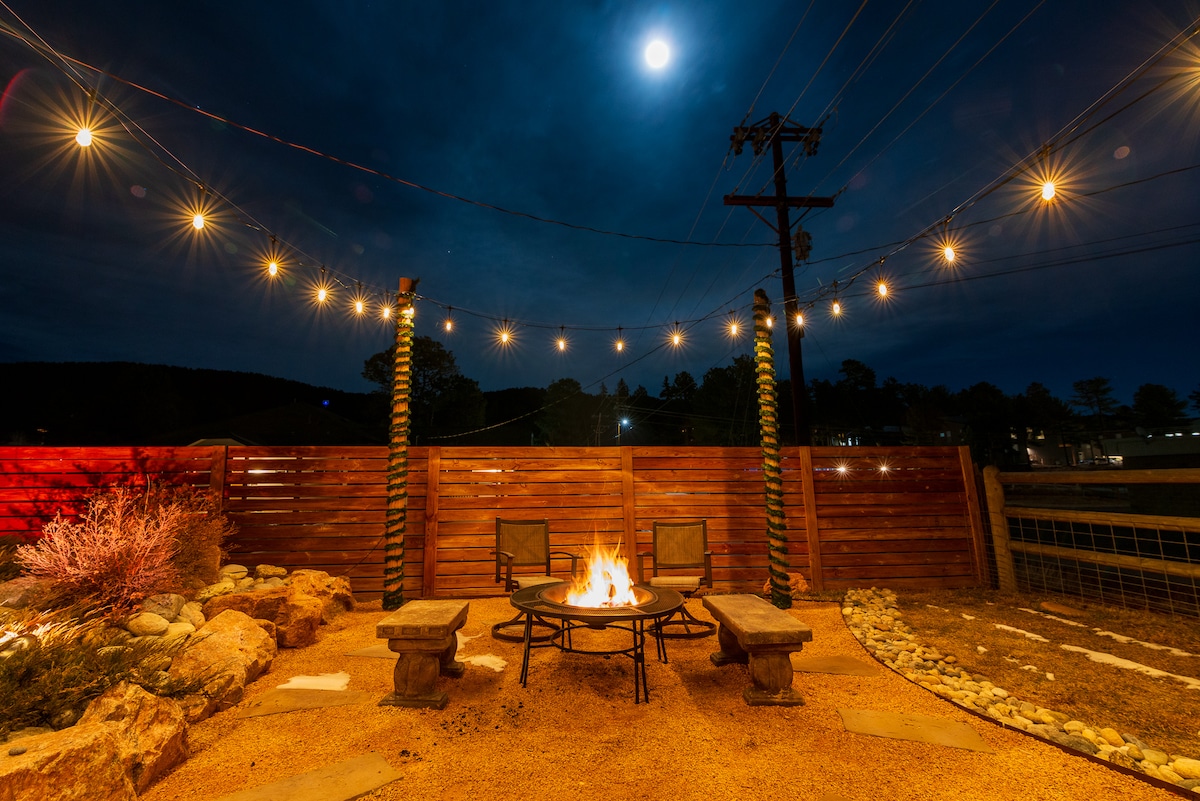 An outdoor fire pit area features comfortable seating arranged around a central fire. Soft glowing string lights stretch overhead, enhancing the inviting ambiance. Natural stones and gravel surround the space, while a wooden privacy fence provides a sense of seclusion under the night sky.