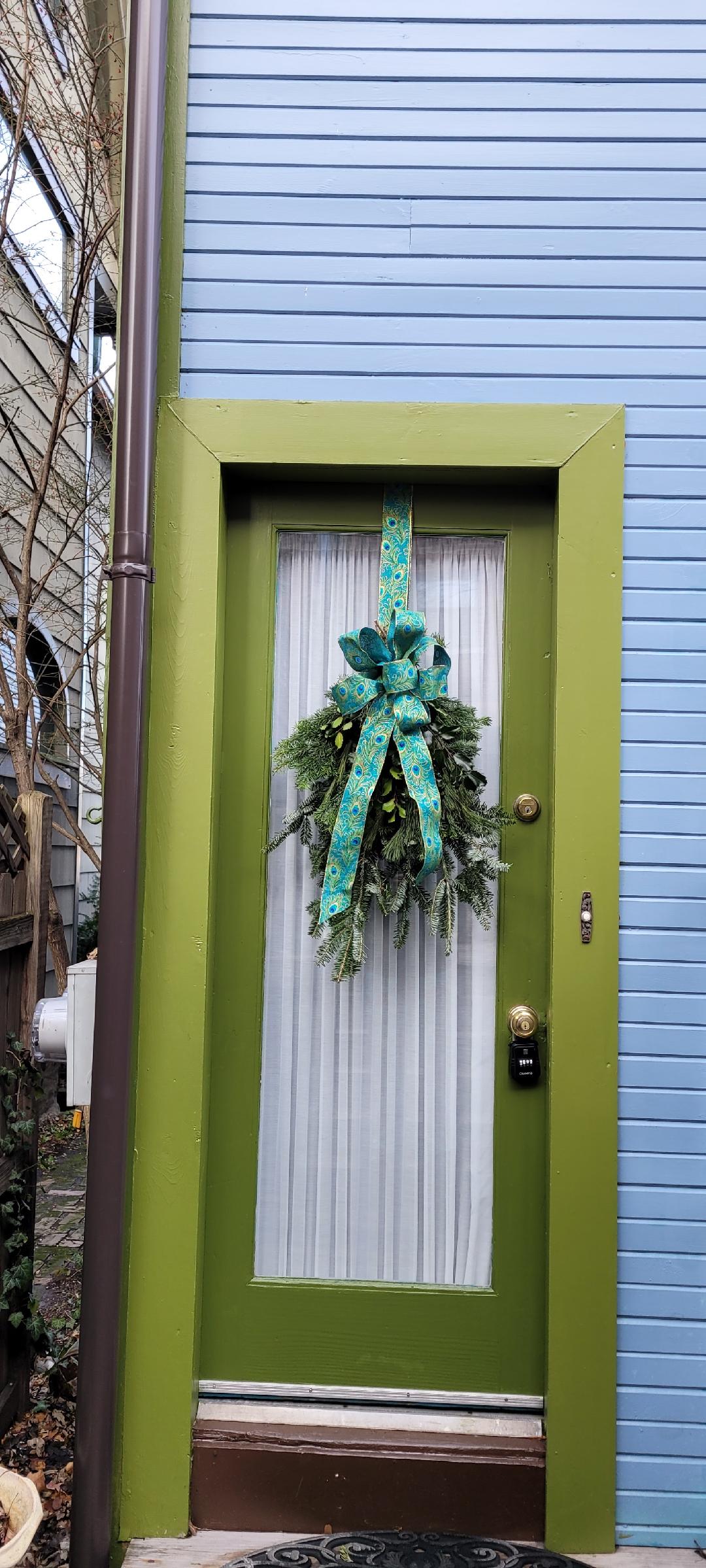 The image features a green door adorned with a festive wreath tied with a bright blue and green bow. A sheer white curtain is visible behind the door, and the wall beside the door is painted in a soft blue shade.