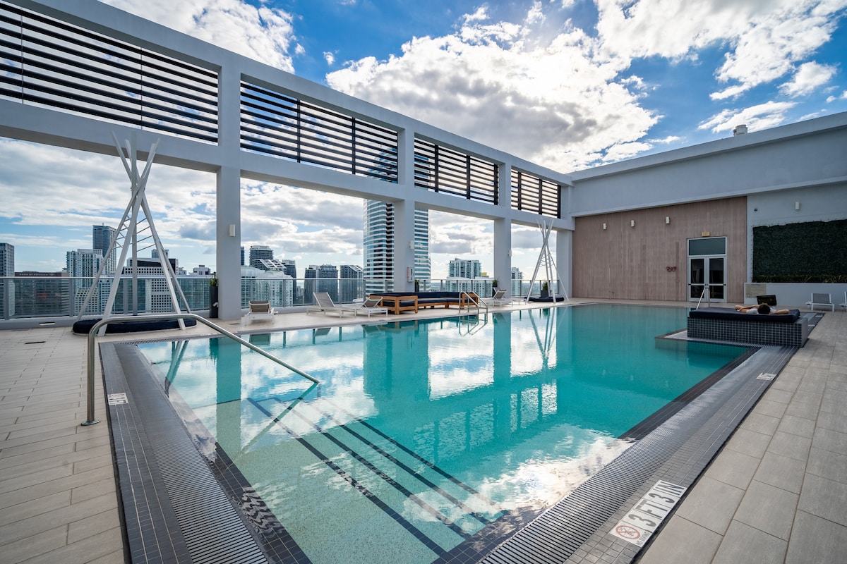 A rooftop pool is visible, surrounded by sleek lounge chairs and urban views. The pool features clear water that reflects the sky, while a seating area includes a sofa and side tables. Large windows create an open, airy atmosphere.