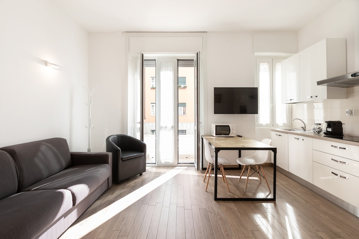 A modern living space features a dark sofa and a black armchair, complemented by a small dining table with white chairs. Natural light fills the room through floor-to-ceiling windows, which lead to a balcony. A wall-mounted TV is visible, enhancing the functional layout.