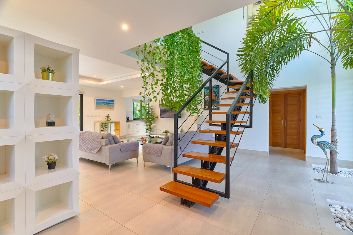 An airy living area is highlighted by a modern staircase with wooden steps and black railings. Lush greenery drapes down from the upper level, enhancing the space. Comfortable gray sofas are positioned nearby, creating an inviting setting for relaxation. Natural light floods in through large windows.