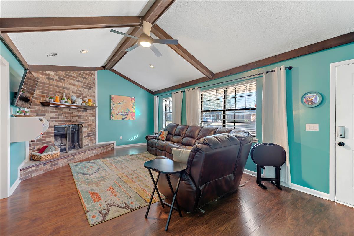 A spacious living room is presented, featuring a comfortable leather sofa and a patterned area rug. The room is highlighted by vaulted ceilings and wood beams. A flat-screen TV is mounted on the wall, and a decorative fireplace adds a cozy focal point.