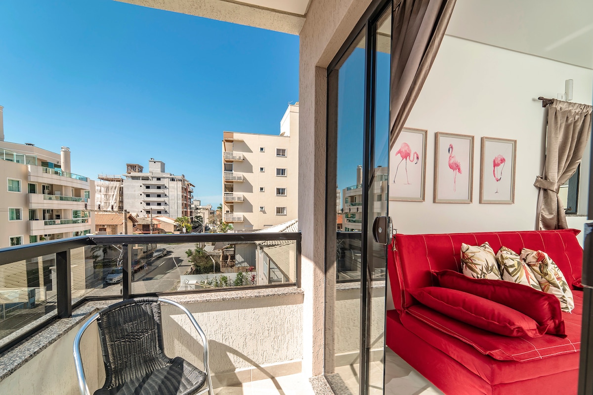 A balcony offers a view of the surrounding area, featuring modern buildings and clear skies. A black chair is positioned on the balcony, while a red sofa adorned with decorative pillows is visible in the adjacent living area, with light streaming through the glass doors.