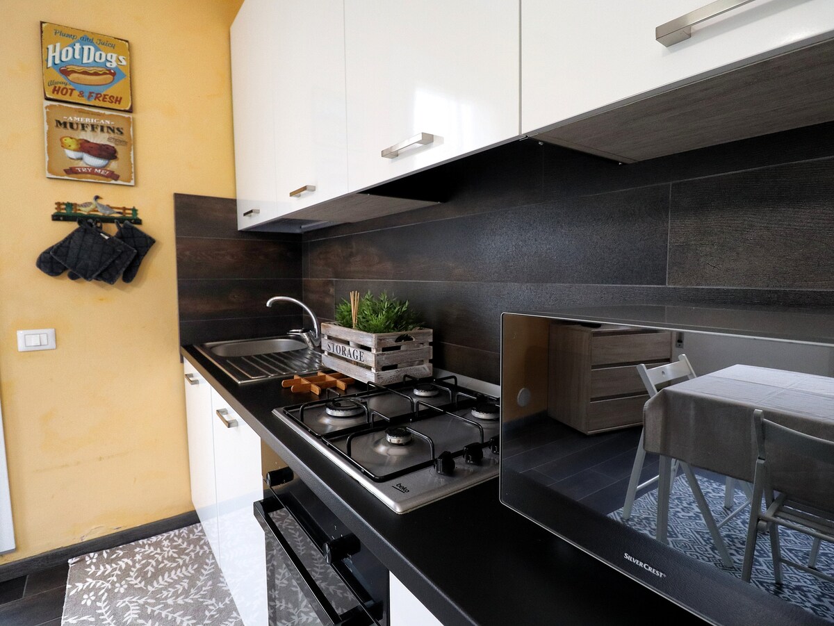 The kitchen features a modern design with white cabinetry and dark wood accents. A gas stovetop is visible alongside a microwave and a stainless-steel sink. Decorative elements include a small plant in a wooden box and hanging kitchen towels.