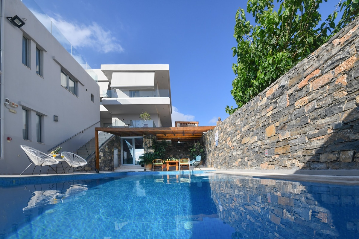 A shimmering pool reflects the blue sky, surrounded by modern stone walls and greenery. The outdoor area includes lounge chairs and a shaded seating space, creating an inviting environment for relaxation near the building's contemporary architecture.