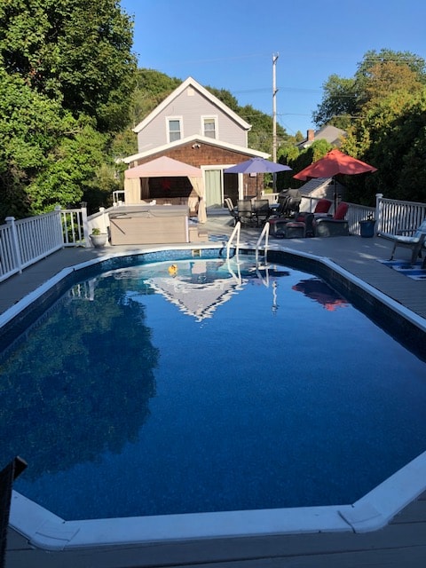 An inviting outdoor pool glistens under clear blue skies, with surrounding wooden deck areas featuring multiple shaded seating options. The cottage is visible in the background, surrounded by greenery. A bright umbrella adds a pop of color to the relaxing space.