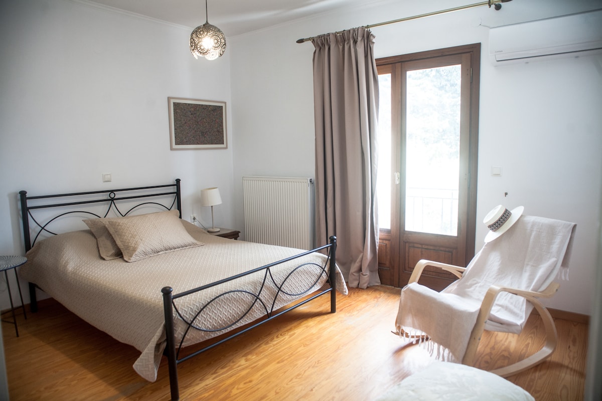 A serene bedroom features a spacious bed with a neutral-toned covering. A comfortable chair is positioned next to a window, allowing natural light to enter the room. A simple lamp and a decorative artwork add subtle charm, while curtains frame the glass doors leading to a balcony.