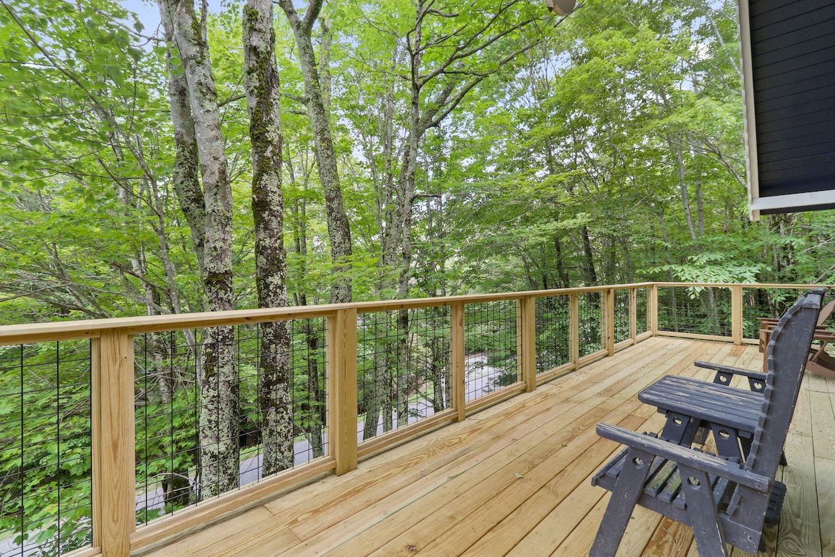 A wooden deck is surrounded by lush trees, offering a serene outdoor space. The deck features a railing with a wire design, and two dark chairs are positioned for relaxation. The natural light filters through the leaves, creating a calming atmosphere.