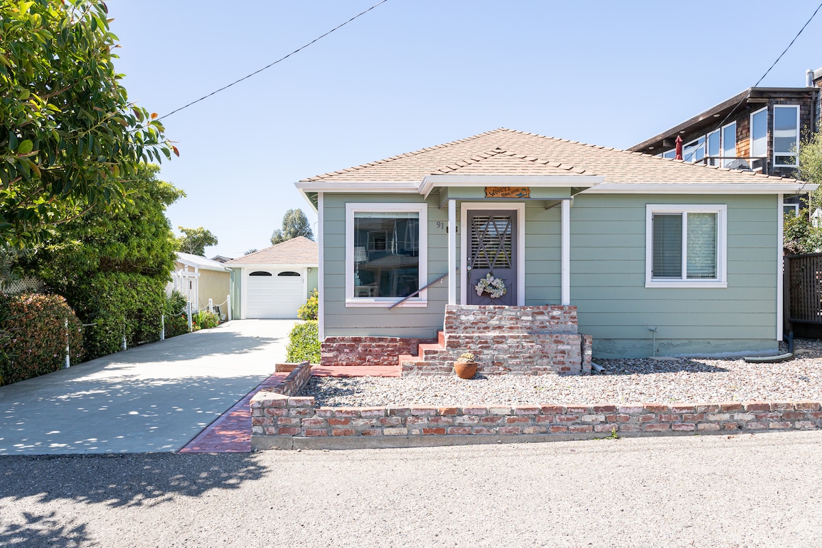 The exterior of a charming beach cottage is captured, featuring light green siding, a welcoming front porch, and a decorative entrance. A gravel yard with a few potted plants is visible, along with a partially paved driveway leading to a garage in the background.