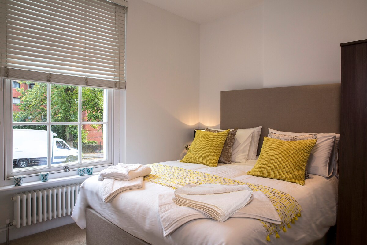 A well-lit bedroom features a spacious bed adorned with decorative yellow cushions and a textured throw. Large windows allow natural light to illuminate the space, with partial views of greenery outside. A classic wardrobe stands against one wall, adding to the room’s functional design.