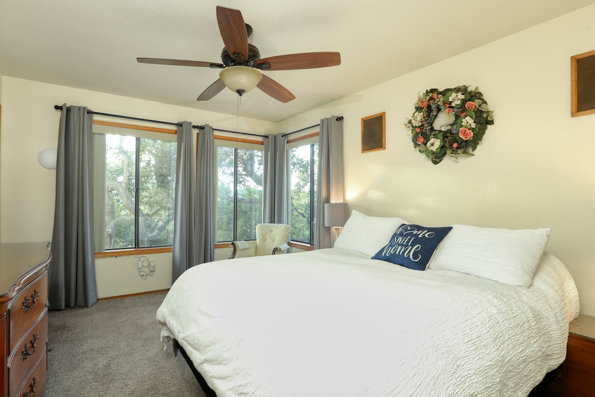 A well-appointed bedroom features a spacious bed with crisp white bedding, set against a neutral wall. Natural light filters through large windows adorned with light gray curtains, revealing a view of oak trees. A ceiling fan enhances the room's comfort.