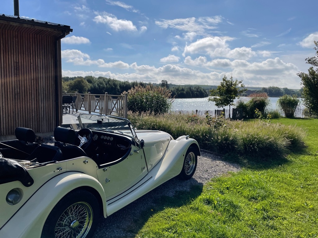 A classic convertible car is parked on a gravel drive beside a lush green lawn. In the background, a serene lake is visible, surrounded by vibrant foliage under a clear blue sky with fluffy white clouds.