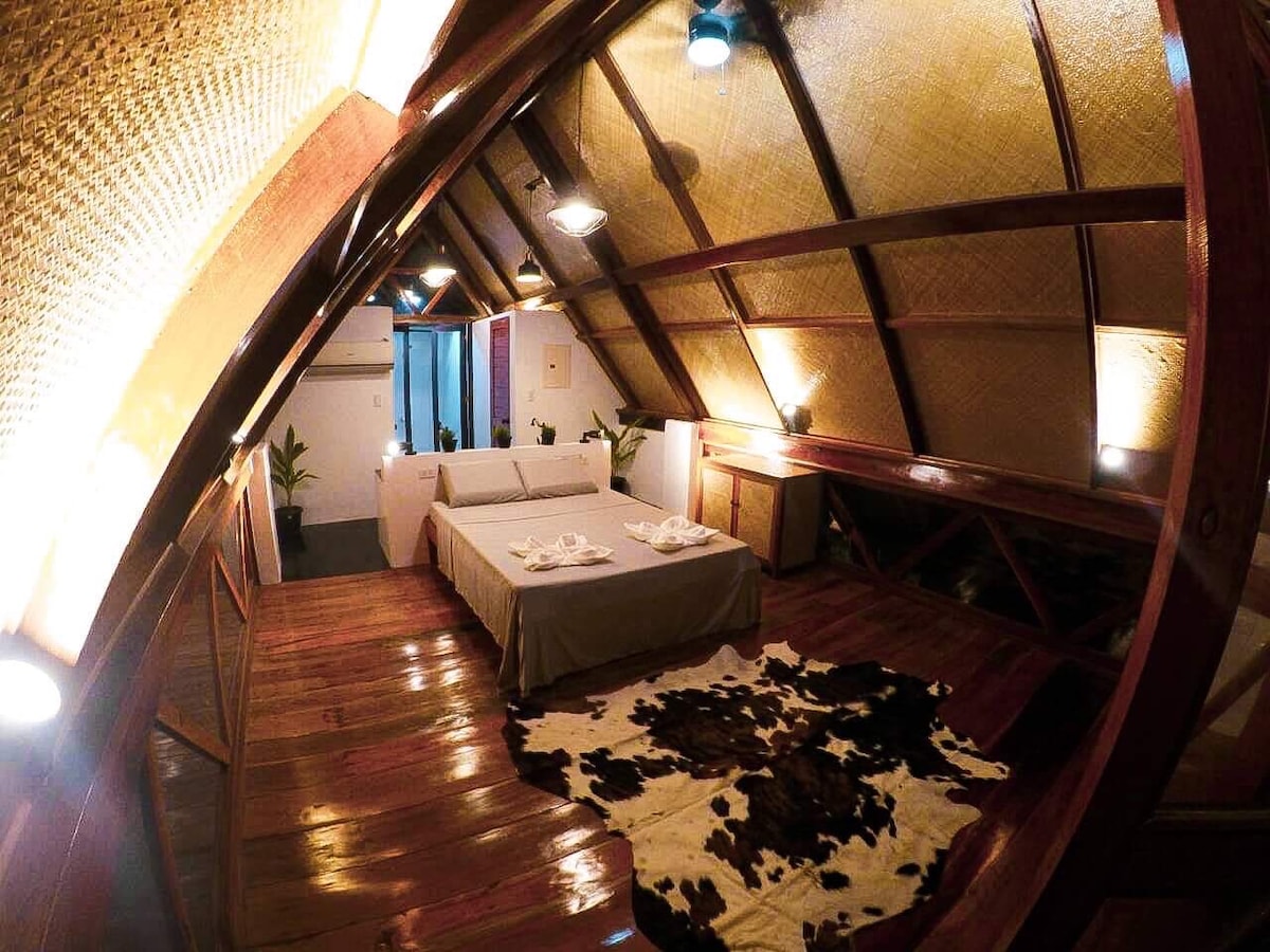 A cozy sleeping area features a spacious bed dressed with neutral linens. A cowhide rug lies on the polished wooden floor. Soft lighting emanates from ceiling lights, illuminating the bamboo ceiling and showcasing greenery in the background.
