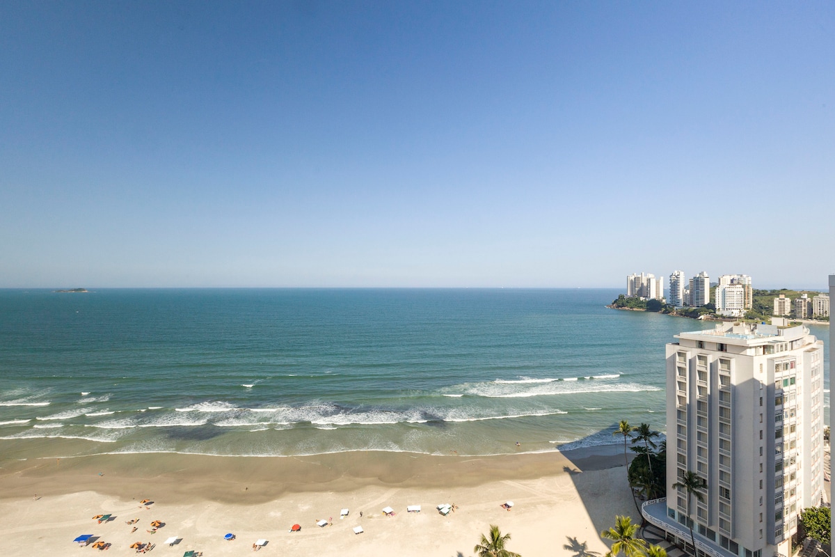 A wide ocean view stretches across the frame, revealing sandy shores and gentle waves. A few colorful beach umbrellas dot the shoreline, while modern buildings are visible in the distance under a clear blue sky.