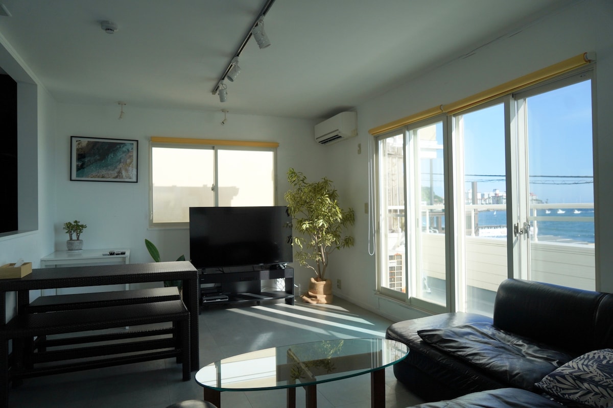A bright living area is highlighted by large windows offering views of the sea and allowing natural light to fill the space. A comfortable dark leather sofa faces a flat-screen TV, with a glass coffee table in front. A potted plant adds a touch of greenery.