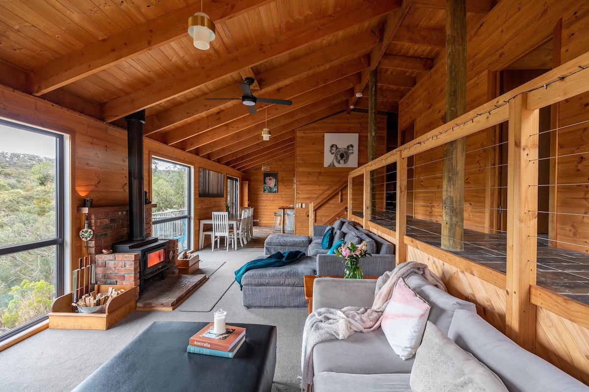 A spacious living area features wooden beams and floor, providing a warm atmosphere. Comfortable seating arrangements include sofas and an ottoman. A cozy fireplace serves as a focal point, while large windows present views of the surrounding natural landscape.