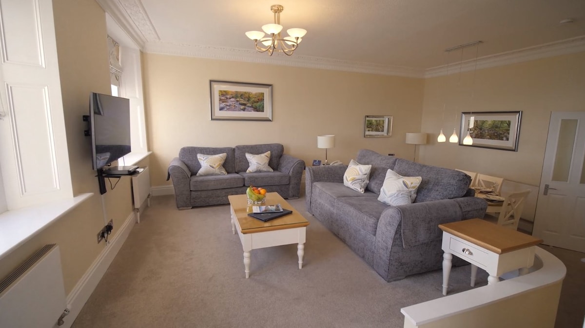 A spacious living area is furnished with two comfortable sofas and a light-colored armchair. A flat-screen television is mounted on the wall, while soft lighting is provided by a chandelier and pendant lights. A small coffee table sits in the center, accented by a tray with fruit.