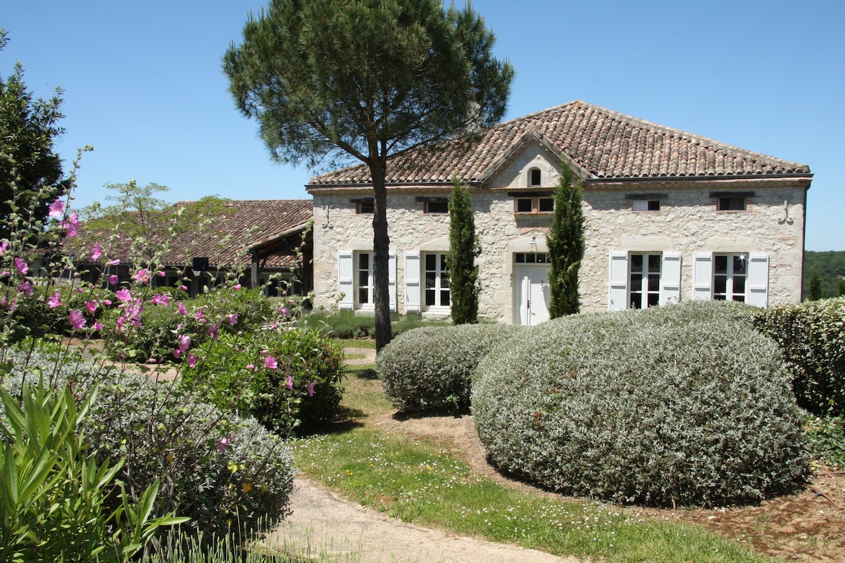 A charming white stone house is surrounded by lush gardens, featuring neatly shaped shrubs and vibrant flowers. The roof is adorned with traditional tiles, and large windows invite natural light, enhancing the inviting facade. A shaded area is visible near the entrance.