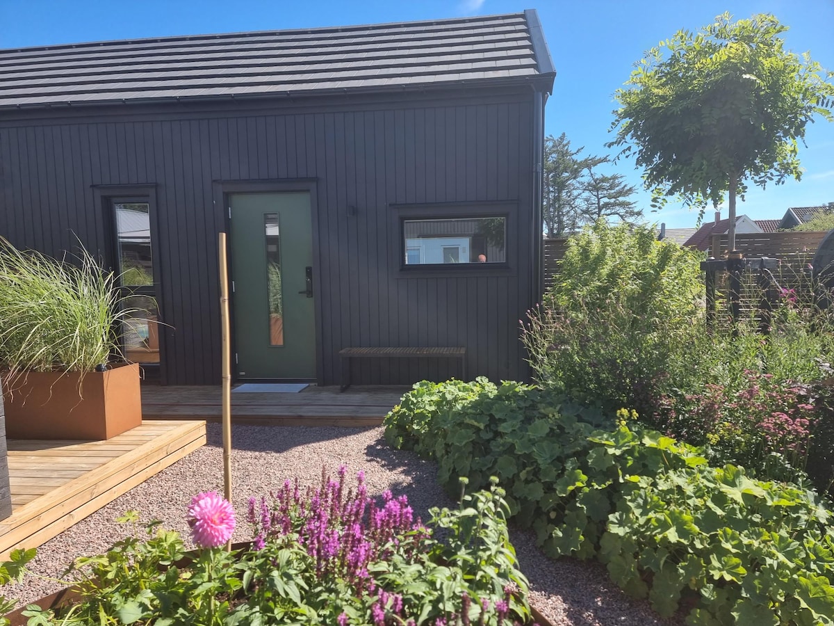 The entrance of the guesthouse is framed by lush, colorful plants, including blooming flowers. The dark wood exterior is complemented by a green front door and large window, inviting light into the space. A wooden bench sits beside the door on a well-maintained deck.