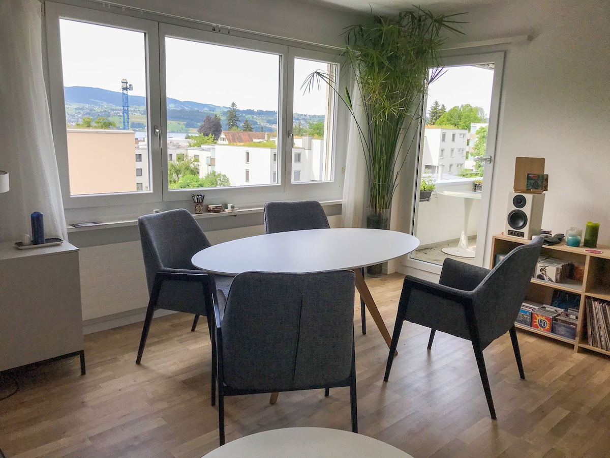 A dining area is presented, featuring a round white table surrounded by four gray upholstered chairs. Large windows allow for natural light and offer views of green hills and nearby buildings. A potted plant adds a touch of nature to the space.