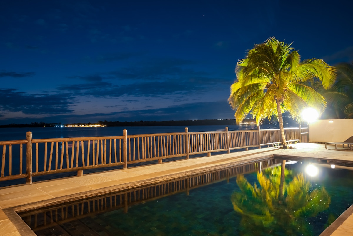 The serene outdoor area features a swimming pool reflects the night sky. Palm trees stand gracefully nearby, and the illuminated deck provides a gentle glow against the water. A wooden railing borders the space, enhancing the connection with the beachfront scenery.