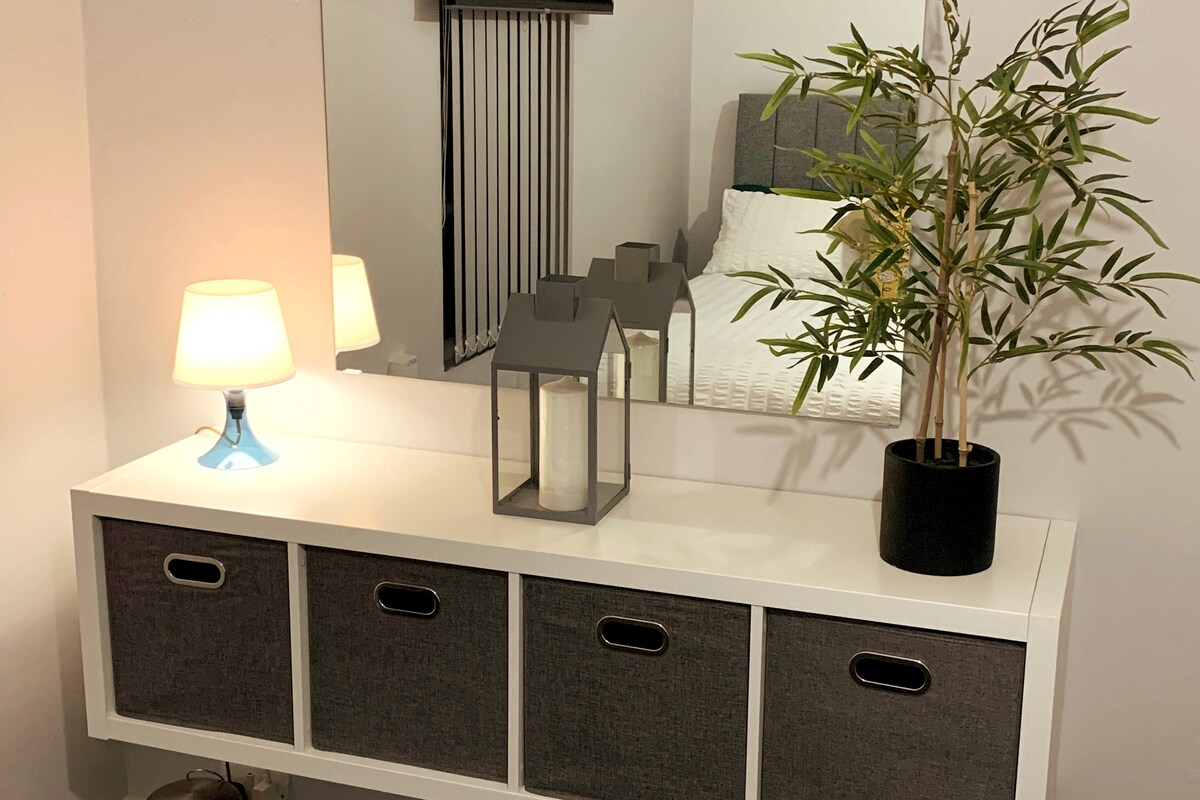 A sleek console table is seen against a neutral wall, featuring storage drawers with grey fabric fronts. A table lamp casts a soft glow beside a decorative lantern, while a potted plant adds a touch of greenery. A mirrored surface reflects the nearby bed.