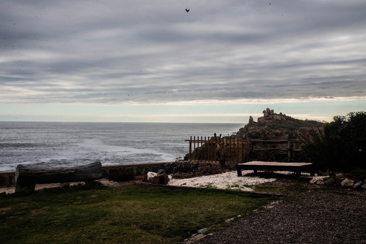 A coastal view captures the ocean's waves gently lapping against the rocky shore. A grassy area is framed by wooden fencing, with scattered stones and a weathered log positioned prominently. The sky is filled with overcast clouds, creating a serene atmosphere.