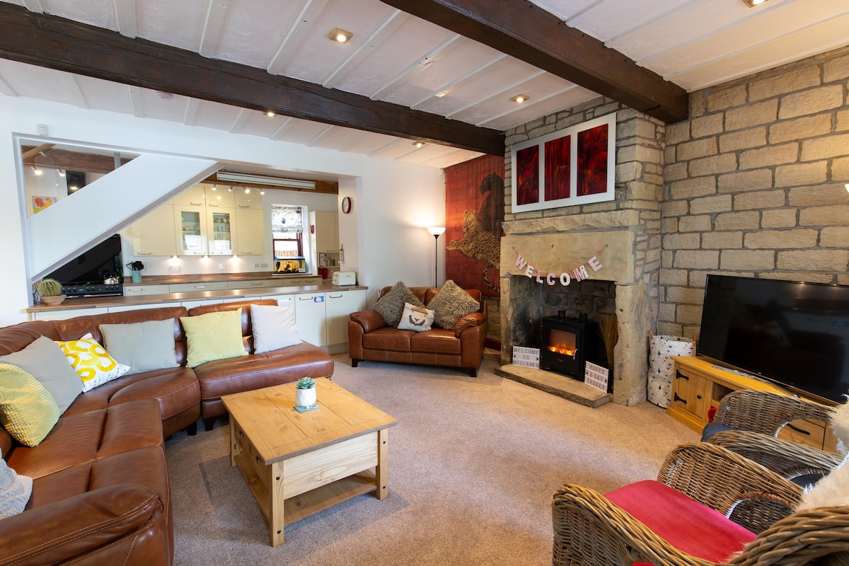 The spacious lounge area features a comfortable leather sectional sofa and a wooden coffee table. A stone fireplace with an electric stove adds warmth, while a large TV is positioned nearby. Light from large windows illuminates the room, enhancing the welcoming feeling.