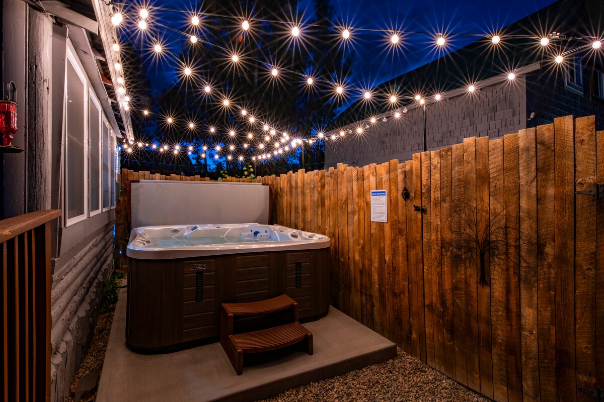 A private hot tub is surrounded by a wooden fence, illuminated by string lights overhead, creating a cozy atmosphere in the evening. Steps lead up to the hot tub, which is placed on a level concrete surface among gravel.