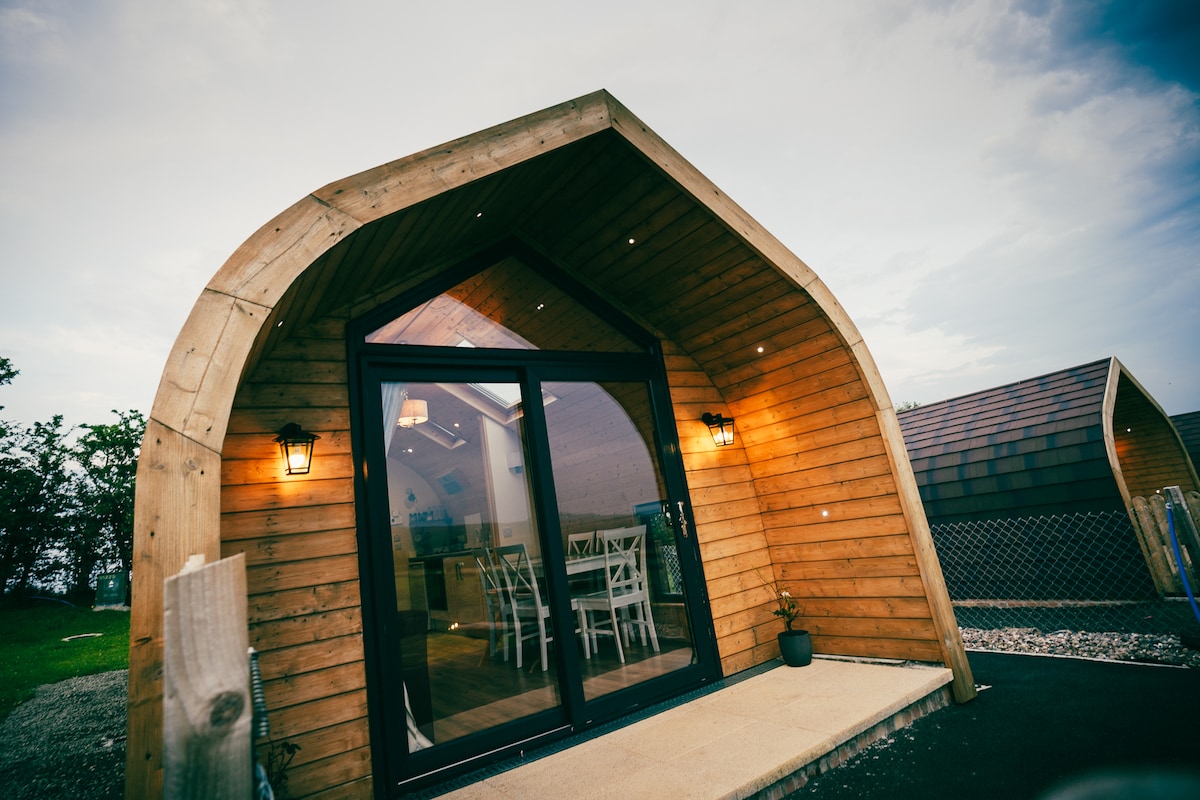 The entrance of a glamping cabin is showcased, featuring a distinctive arched design and wooden exterior. Large glass doors provide a view into a bright interior, while outdoor lighting adds a welcoming touch. A small potted plant sits beside the entrance.