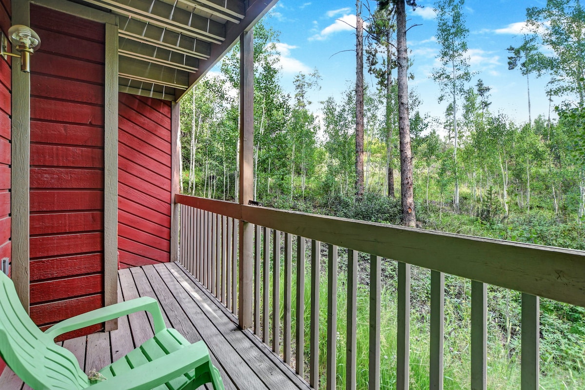 A covered balcony features two green chairs positioned for relaxation, overlooking a serene landscape of trees and foliage. The wooden railing and floorboards add a natural touch, with glimpses of distant greenery visible beyond the balcony.