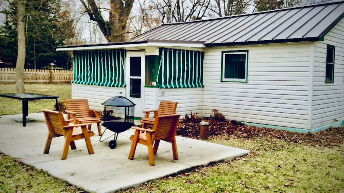 A rustic cottage is shown with a metal roof and green striped awnings. Four wooden chairs surround a charcoal grill on a patio, offering a comfortable outdoor space. The cottage's exterior is surrounded by grass and is partially shaded by trees.