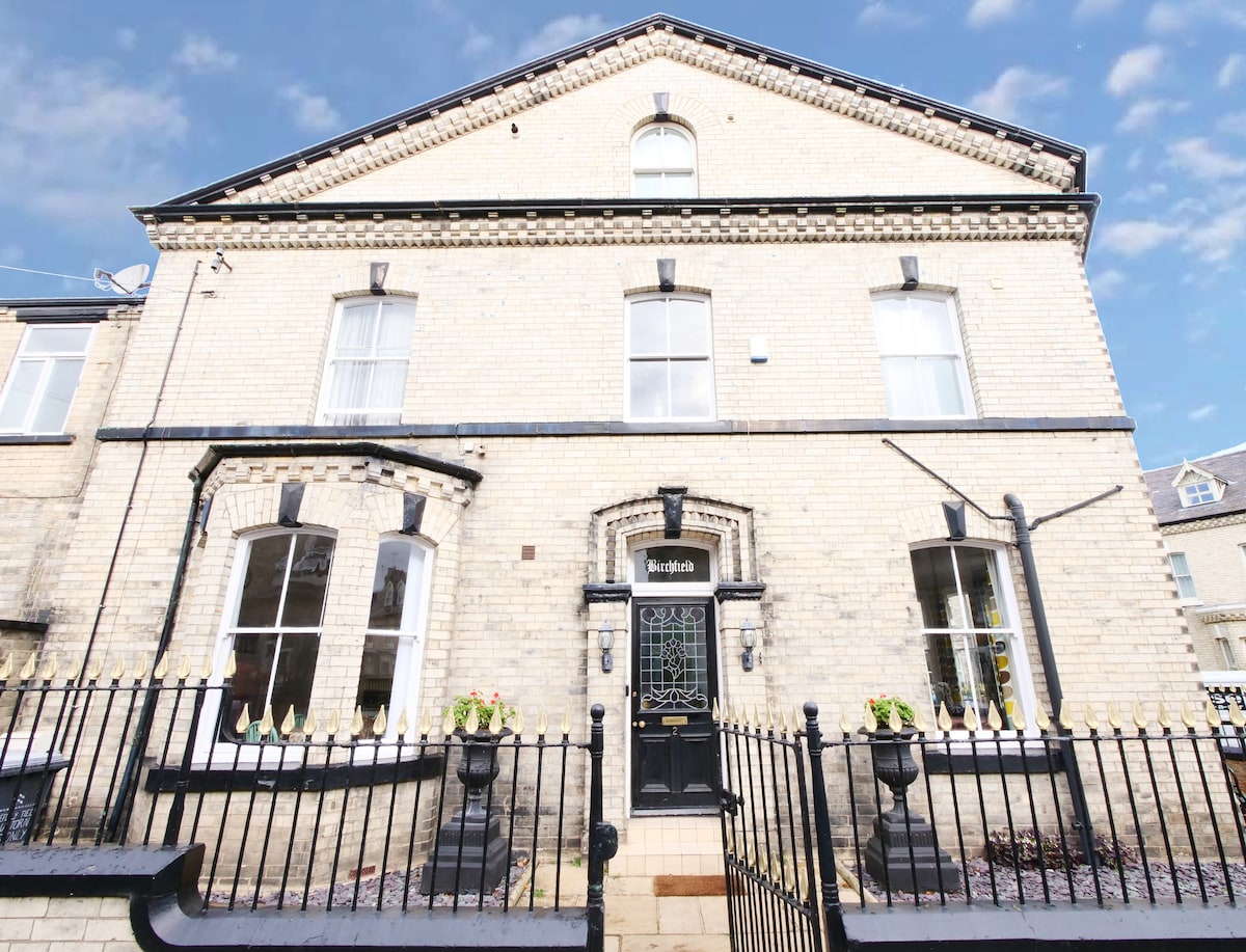 The exterior of Birchfield, a large Victorian house, is displayed, characterized by its elegant brick facade and prominent windows. The entrance features a decorative door framed by stone accents. A wrought-iron fence with planters adds to the appeal, inviting guests inside.