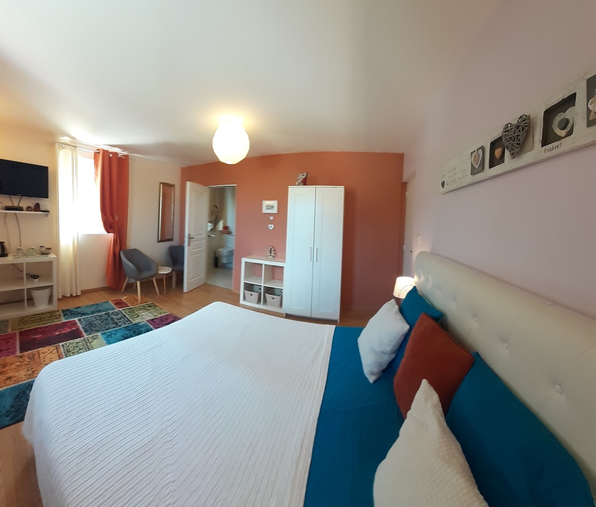 A well-lit bedroom features a spacious bed with a blue and white bedspread. A television is mounted on the wall. A closet and a small seating area with two chairs are visible. Colorful rugs add visual interest to the wooden floor.
