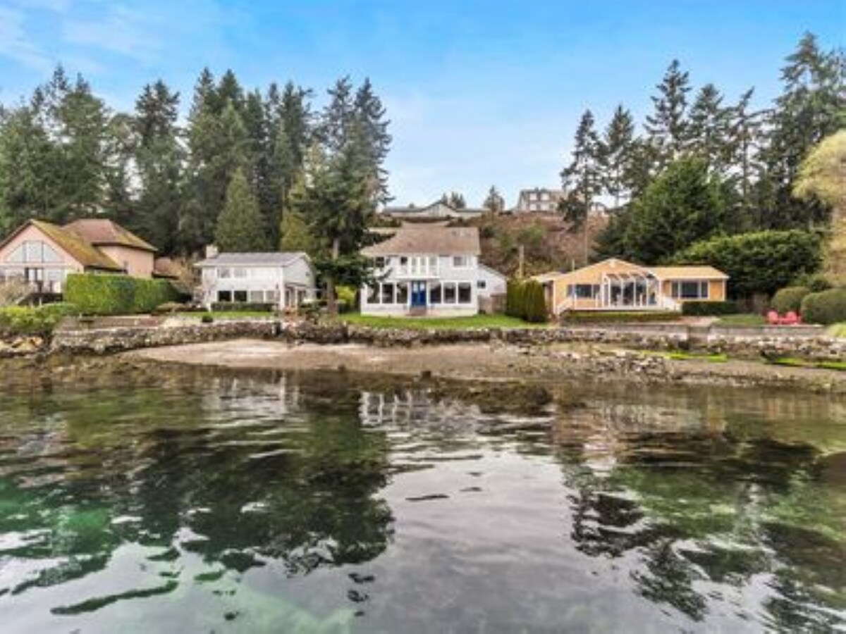 A waterfront view showcases several private homes surrounded by lush trees. The clear water reflects the natural landscape, and a beach area is visible in the foreground. Various seating arrangements can be seen along the shoreline, inviting relaxation by the water.