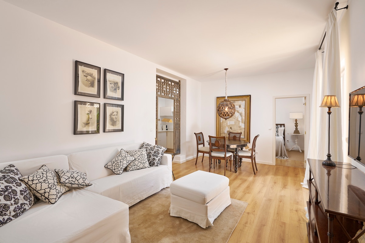 A bright living area features a white sofa adorned with patterned cushions, complemented by a round ottoman. Four wooden dining chairs surround a rectangular table, while framed artwork adorns the walls. Natural light fills the space through large windows, enhancing the warm wood flooring.