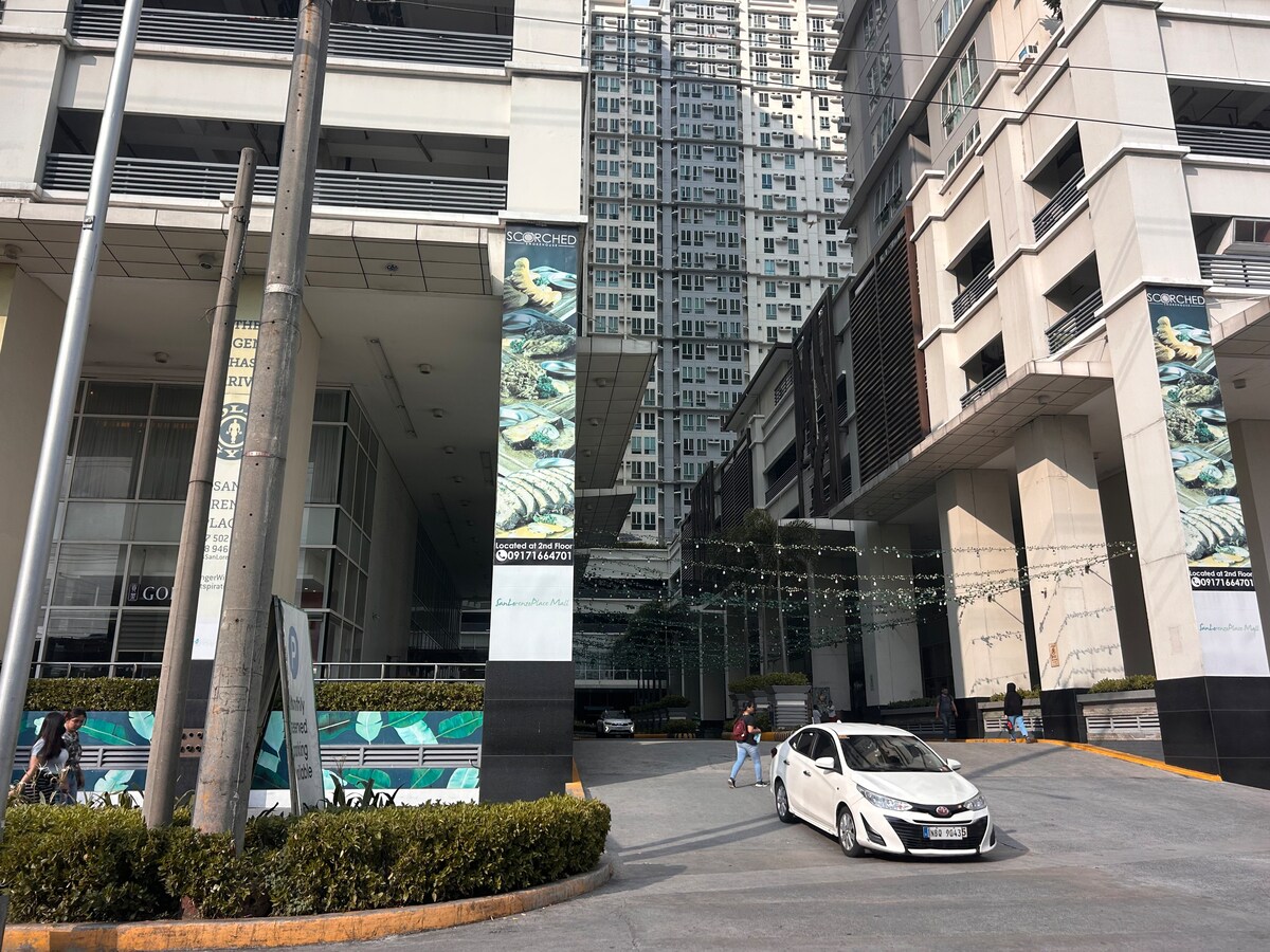 The image depicts a modern high-rise building with a sleek design. The entrance is characterized by a spacious driveway lined with greenery. A white vehicle is parked, and large banners are visible on the building's exterior. Tall residential towers rise in the background.