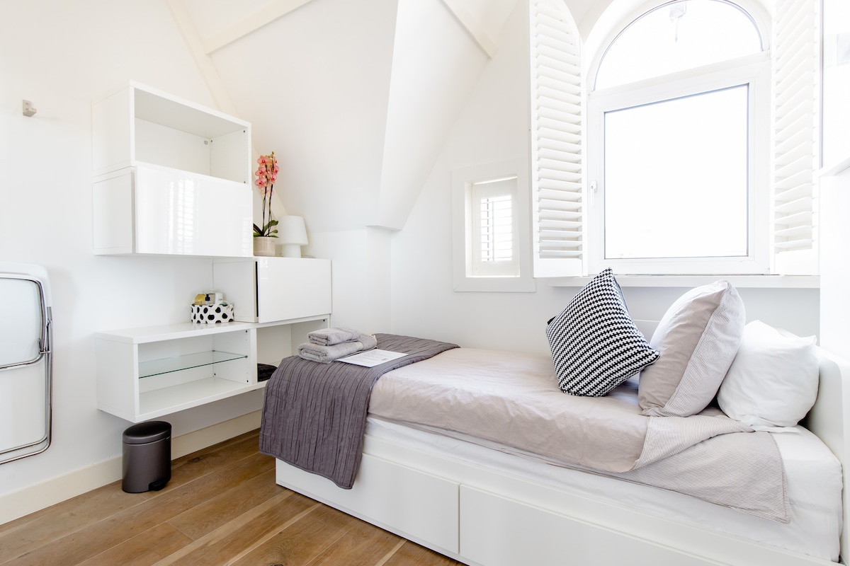 A bright bedroom features a single bed with neutral bedding and decorative pillows. A small shelf with a plant and decor sits against the wall. Light enters through a window with white shutters, complementing the wooden floor and minimalist design.