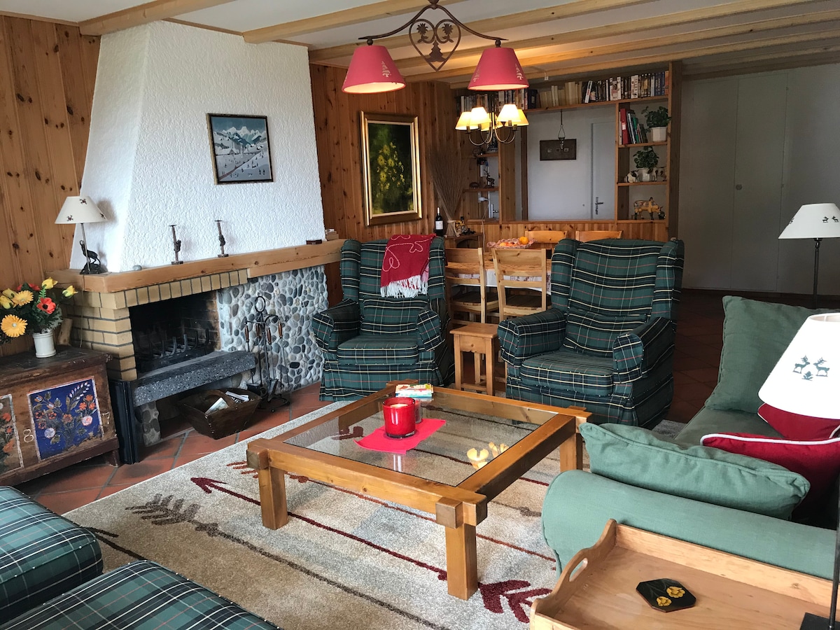 A comfortable living area is featured with wooden paneling and a stone fireplace. Two plaid armchairs and a green sofa are arranged around a wooden coffee table, holding red candles and snacks. Shelves with books are visible in the background, adding functionality.